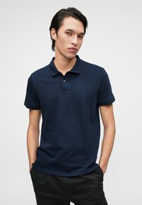 Sötétkék pólóing, pamutból készült, három gombos nyakkivágással és egy kis hímzett logóval a mellrészen. Rövid ujjak, sima textúra.