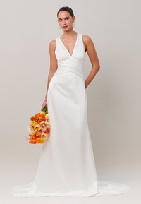 Femme portant une robe de mariée blanche en satin sans manches avec un décolleté en V profond, tenant un bouquet de fleurs orange et roses.