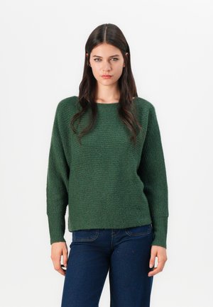 Grønn strikkegenser med løs passform, lange ermer og teksturert overflate. Kombinert med mørke jeans for en avslappet stil.