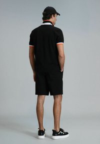 Zwart poloshirt met contrasterende witte en oranje kraag, gecombineerd met zwarte shorts en zwarte sneakers met witte zolen. Geplaatst tegen een neutrale achtergrond.