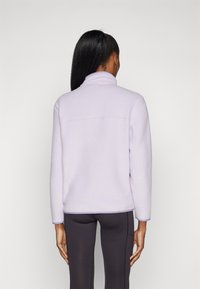 Lila fleece jas met hoge kraag, raglanmouwen en contrasterende afwerking, gecombineerd met nauwsluitende donkere leggings. Zachte textuur, casual pasvorm.