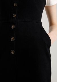 Svart denim_jumpdräkt med åtsittande överdel, rund halsringning och knäppning framtill. Har sidofickor och en len textur.