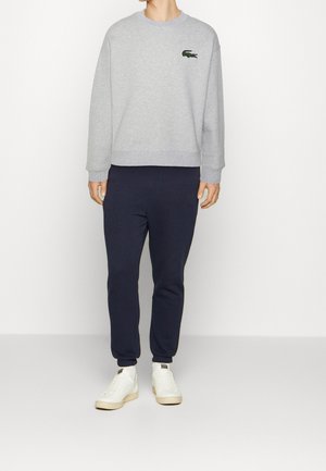 Grå tröja med grön broderad logotyp, kombinerad med marinblå mjukisbyxor. Modellen bär vita sneakers. Avslappnad klädsel, mjukt tyg, ledig passform.