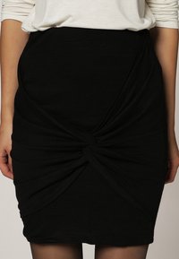 Svart åtsittande kjol gjord av ett texturerat, elastiskt tyg, med ett vridet detalj framtill och en knälång design.