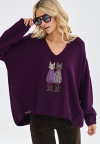 Fioletowy sweter w serek z haftem przedstawiającym dwa koty w płaszczach, jeden w fioletowe kropki, a drugi w wzorzysty design. Luźny krój.