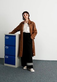 Casaco trench de couro castanho sobre um polo branco com um logotipo, calças largas pretas com riscas brancas e ténis brancos. Armário de arquivos azul ao lado.