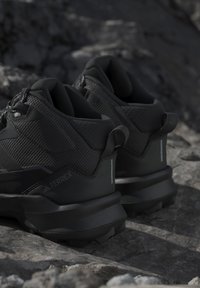 Sorte Adidas Terrex vandrestøvler med strukturerede sider og tykke såler, placeret på klippefyldt underlag udendørs.