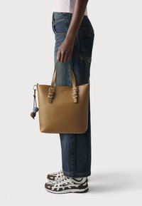Person wearing blue jeans and white-brown sneakers holding a tan leather tote bag with two handles and decorative hanging balls.