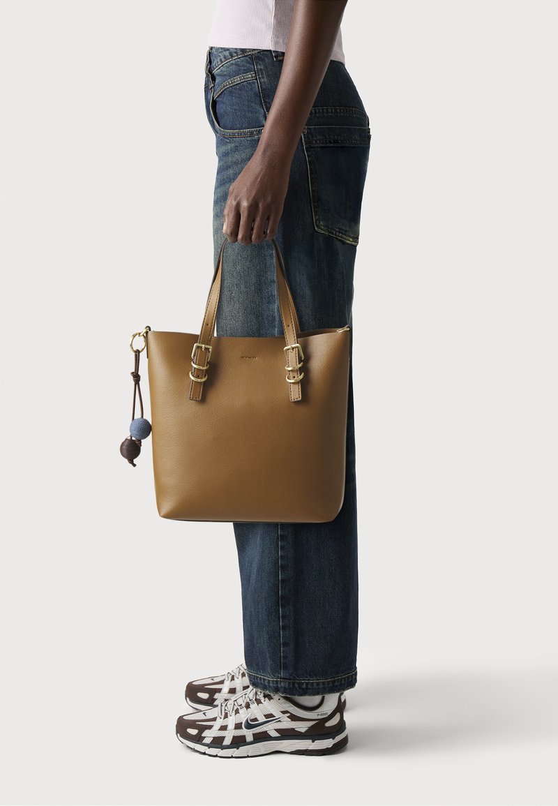 Person wearing blue jeans and white-brown sneakers holding a tan leather tote bag with two handles and decorative hanging balls.