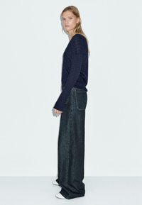 Marineblaue gerippte Strickjacke mit langen Ärmeln, kombiniert mit hoch taillierten, weit geschnittenen, dunklen Jeans und weißen Sneakers, vor einem schlichten Hintergrund posiert.