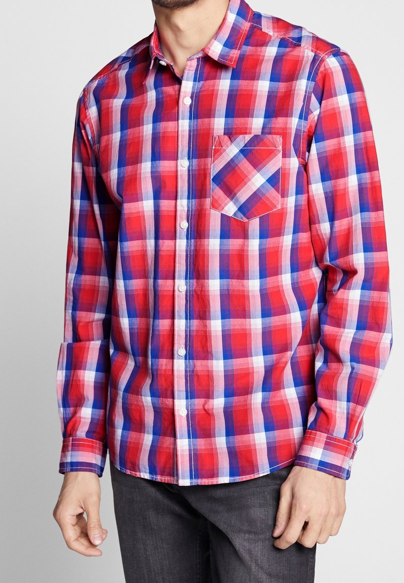 Man i rutig skjorta i röd, blå och vit färg med lång ärm och knäppning framtill, med en framficka och mörka jeans, står mot en enfärgad bakgrund.