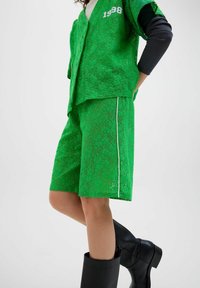 Zielona koszula z krótkim rękawem i dopasowane krótkie spodenki wykonane z teksturowanej koronki, z białym paskiem na spodenkach i haftem z napisem „1998”. Czarne kozaki za kolano.