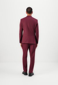 Bordeaux pak met een getailleerde pasvorm, voorzien van een enkele vent en bijpassende broek, gecombineerd met zwarte schoenen, gezien vanuit de rug.