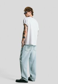Witte katoenen t-shirt met korte mouwen, lichtblauwe baggy jeans, zwarte instappers en zonnebril. Opvallende tatoeages op de onderarmen, ontspannen pasvorm.