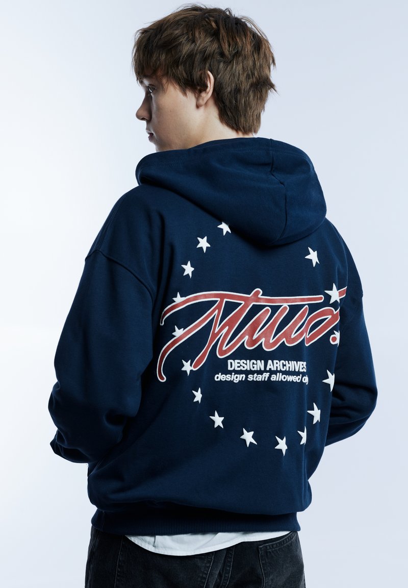 Marineblauer Kapuzenpullover mit großem rotem Schriftzug und weißen Sternen auf der Rückseite, mit Kordelzug an der Kapuze und gerippten Bündchen.