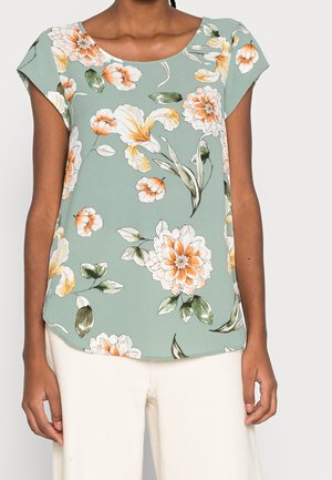 Woman wearing a short-sleeve green blouse with orange and white floral print, paired with white pants.