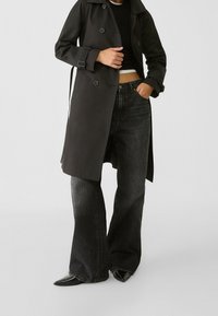 Czarny trenchcoat z podwójnymi guzikami, szerokimi klapami i paskiem w talii, połączony z wysokim stanem i rozszerzanymi ciemnymi dżinsami oraz spiczastymi butami.