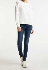 White sweatshirt with crew neckline and embroidered black umbrella logo, paired with dark blue skinny jeans and white sneakers with black stripes.