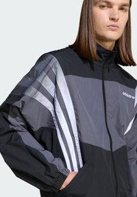 Čierno-sivá bunda Adidas so zipsom vpredu, vysokým golierom a tromi bielymi zvislými pruhmi na pleciach. Ľahká tkanina, voľný strih.