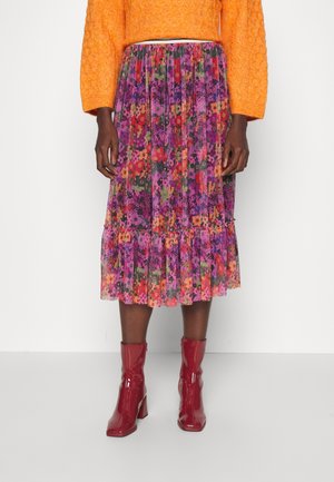 Personne portant une jupe midi plissée à fleurs éclatantes avec des fleurs violettes, rouges et orange, associée à un pull orange vif et des bottines rouges.
