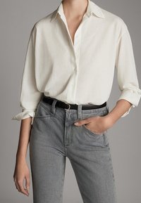 Vit skjorta med uppvikta ärmar, kombinerad med grå jeans och ett svart bälte. Har en klassisk krage och texturerad tyg.