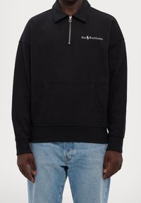 Svart fleece-tröja med dragkedja i kragen, sidofickor och "Polo Ralph Lauren"-logotyp i vitt. Mjuk, varm textur.