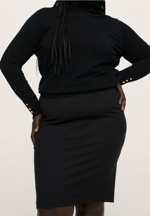 Frau trägt einen schwarzen Rollkragenpullover mit langen Ärmeln und einen figurbetonten schwarzen knielangen Rock, steht mit den Händen in der Hüfte.