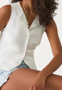 Witte mouwloze blouse met knoopsluiting, textuurstof, gebogen halslijn en subtiele stiksel details, gecombineerd met lichte denim shorts.