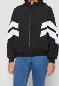 Chaqueta bomber negra con capucha, rayas diagonales blancas en las mangas, cierre de cremallera en el frente, puños elásticos y tejido texturizado. Usada con jeans azules.