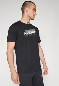 Must mustaheline T-särk mustade lühikeste varrukatega, millel on valge ja hall "HUMMEL" logo, ümar kaelus, valmistatud pehmest kangast, koos tumedate spordipükstega.