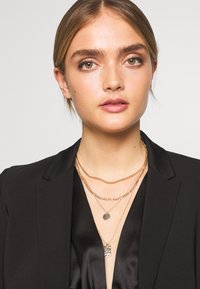 Collares en capas de oro con diseños de eslabones variados y un colgante, llevados sobre un blazer negro y una parte superior de satén, destacando texturas y contrastes.