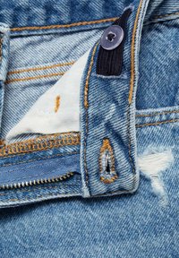 Denim de culoare bleu deschis cu zone uzate, cusături portocalii și închidere cu fermoar. Include o etichetă din material textil și detalii cu nasture.