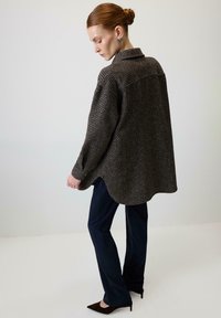 Brązowa, oversize koszula o wzorze jodełki z mankietami zapinanymi na guziki, w zestawieniu z ciemnoniebieskimi spodniami i czarnymi, spiczastymi butami.