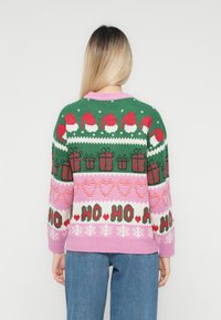 Kvinde iført en farverig julesweater med julehuer, gaver, hjerter, snefnug og "HO HO HO" mønstre, set fra ryggen.