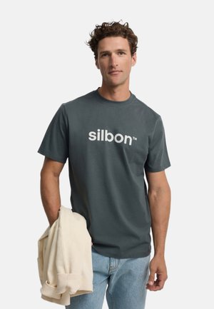 Hombre con camiseta "silbon" gris oscuro sosteniendo una chaqueta beige clara en una mano, de pie frente a un fondo liso.
