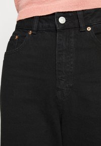Černé džíny z denimu s hladkým povrchem, designem s pěti kapsami, měděnými nýty a předním zapínáním na knoflík. Pás je viditelný nad růžovým svetr.