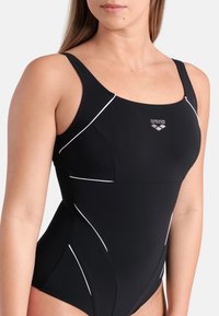 Mulher vestindo um fato de banho preto de uma peça com detalhes brancos e o logótipo "arena" no peito, mostrada dos ombros aos quadris.