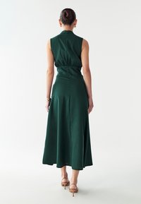 Grünes, ärmelloses Kleid mit enger Taille und fließendem Rock. Hergestellt aus strukturiertem Stoff, verfügt über einen hohen Kragen und einen Rückenreißverschluss.