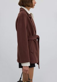 Hnědý oversize blazer ze směsi vlny s páskem v pase, zdobený švy a s klopovým límcem, nošený přes pruhovanou košili a černé šortky.