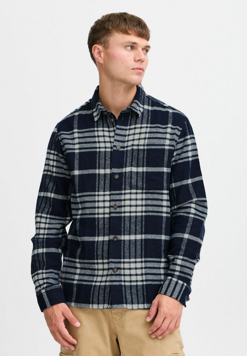 Granatowa i szara koszula flanelowa w kratę z kołnierzykiem button-down, długimi rękawami i kieszenią na piersi. Posiada brązowe zapięcia na guziki oraz miękką fakturę.