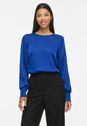 Femme aux longs cheveux foncés portant un chemisier bleu vif à manches longues avec des poignets smockés et un pantalon noir taille haute, souriante à la caméra.