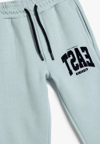Pantalones de chándal azul claro con cinturilla elástica y cordones negros. Presenta un logotipo negro "EAST" impreso en la pierna izquierda.