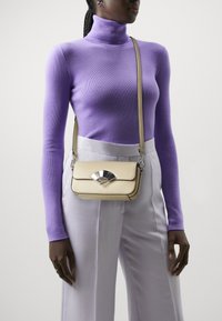 Borsa a tracolla beige in pelle con una chiusura a forma di ventaglio argentato, di forma rettangolare e tracolla regolabile, indossata su un dolcevita viola.