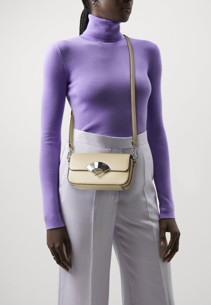 Borsa a tracolla beige in pelle con una chiusura a forma di ventaglio argentato, di forma rettangolare e tracolla regolabile, indossata su un dolcevita viola.