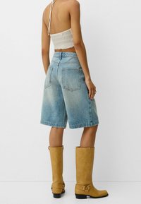 Kobieta w jasnoniebieskich dżinsowych szortach do kolan, beżowym dzianinowym topie na krótki rękaw i wysokich, jasnobrązowych zamszowych butach, stojąca na białym tle.