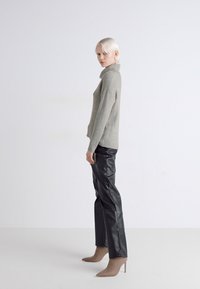 Lichtgrijze geribbelde trui, zwarte neplederen broek, beige haklaarzen tot de enkel; model in profiel tegen een effen achtergrond.