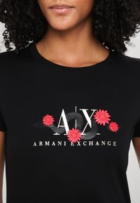 Melna kokvilnas T-kreklu ar baltu "AX" logo un "ARMANI EXCHANGE" tekstu, izrotāts ar sarkanu ziedu izšuvumu un melna čūskas dizainu.