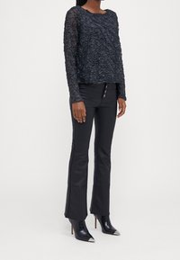 Schwarzes, strukturiertes Langarmoberteil mit einem dekorativen Muster; kombiniert mit engen schwarzen Hosen mit Knopfdetails und spitzen schwarzen High Heels.