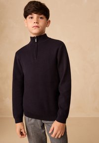 Meresinine tõmblukuga kampsun, millel on kõrge krae ja pikad varrukad, valmistatud tekstuurse lõngaga materjalist; kombineeritud ruuduliste pükstega.