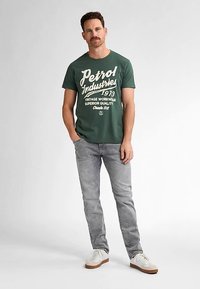 Zelené bavlněné tričko s bílým nápisem ve vintage stylu; doplněné světle šedými džíny a bílými teniskami. Jednoduchý, ležérní střih.